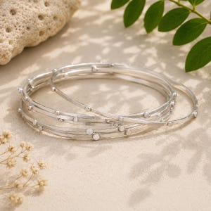 May include: A set of three thin, circular silver bangles. Each bangle features a smooth, polished metallic finish with small, evenly spaced raised silver dots or beads integrated along the outer circumference of the circular bands.