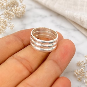 May include: A set of three stacked silver rings with a smooth, polished finish. The rings are circular, metallic, and feature a simple, plain band design. Each ring has a reflective surface that catches the light, creating a clean and uniform appearance.