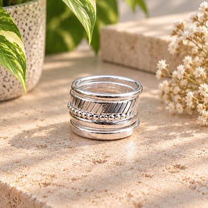 May include: A stack of five silver rings featuring varied textures, including smooth polished bands, a band with diagonal etched lines, and a band composed of small connected spheres, all arranged in a vertical column on a flat surface.