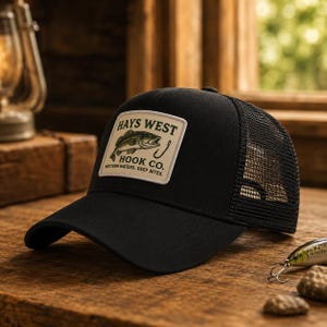 Może przedstawiać: Czarna czapka typu trucker z prostokątną naszywką z przodu. Naszywka przedstawia ilustrację zielonej ryby, haczyk wędkarski oraz napis HAYS WEST HOOK CO. WESTERN WATERS. DEEP BITES. Czapka posiada pełny przedni panel oraz siateczkę z tyłu.