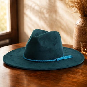 Pode incluir: Um chapéu fedora de aba larga em tom verde-azulado profundo com uma textura lisa e mate. O chapéu apresenta uma copa vincada e uma aba plana. Uma fita de cordão entrançado em turquesa brilhante envolve a base da copa do chapéu.