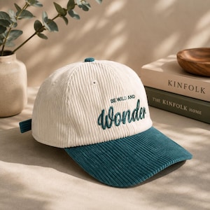 Pode incluir: Um boné de beisebol de veludo cotelê na cor creme com aba verde-azulada escura e o texto bordado "BE WILD AND Wonder" em linha verde-azulada escura no painel frontal. O boné inclui um pequeno botão no topo e uma tira na parte de trás.