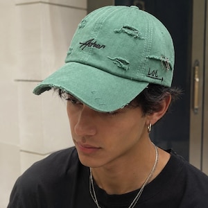 Può includere: Un berretto da baseball verde salvia con una finitura vissuta e usurata, caratterizzato da diversi tagli sfilacciati. Il berretto presenta la scritta ricamata in nero "Asher" sul davanti e "Lol" con una freccia sul lato, arricchito da un anello in metallo argentato.