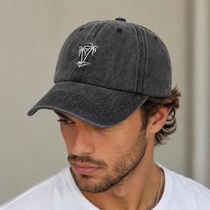 Peut inclure: Une casquette de baseball gris anthracite avec une finition délavée et usée. Le panneau avant présente une petite broderie blanche représentant deux palmiers avec un reflet en dessous. La casquette a une visière courbée et six panneaux.