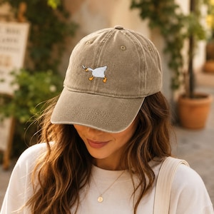 Peut inclure: Une casquette de baseball beige avec une broderie représentant une oie blanche aux pattes orange tenant un petit couteau argenté dans son bec. La casquette présente une visière courbée, une calotte structurée avec des œillets d'aération.