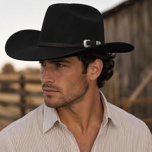 Könnte beinhalten: Ein schwarzer Cowboyhut aus Filz mit einer strukturierten Krone und einer breiten, geschwungenen Krempe. Der Hut ist mit einem dünnen, dunkelbraunen Lederband verziert, das eine dekorative silberfarbene Metallschnalle an der Seite aufweist.