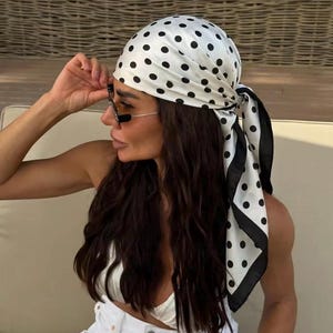 Peut inclure: Un foulard blanc orné d'un motif à pois noirs et d'une bordure noire unie. Ce tissu de forme carrée est noué autour de la tête, avec les extrémités tombant sur le côté, formant un accessoire classique qui recouvre les cheveux et le cuir chevelu.