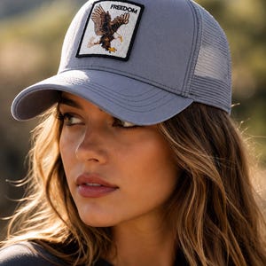 Peut inclure: Une casquette de type trucker grise avec un écusson carré sur le panneau avant. L'écusson présente une illustration d'un aigle brun en vol avec le mot FREEDOM imprimé en lettres capitales noires au-dessus. La casquette a un devant solide et un dos en filet.