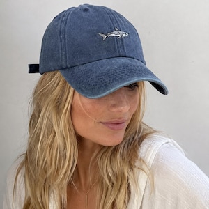Puede incluir: Una gorra de béisbol azul con un acabado desgastado y lavado. El panel frontal presenta un pequeño gráfico bordado detallado de un tiburón en tonos blancos y grises. La gorra tiene una visera curva y una corona estructurada con costuras visibles.