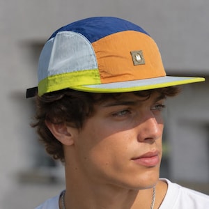 May include: A five-panel cap featuring a colour-blocked design with panels in navy blue, light blue, orange, and lime green. The front panel displays a small, square, olive-coloured patch with a white letter M embroidered in the centre.