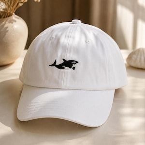 Könnte beinhalten: Eine weiße Baseballkappe mit einer kleinen, aufgestickten schwarz-weißen Orca-Grafik, die mittig auf der Vorderseite platziert ist. Die Kappe verfügt über eine strukturierte Krone, einen gebogenen Schirm und einen passenden weißen Knopf oben.