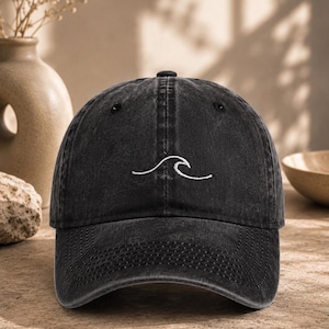 Puede incluir: Una gorra de béisbol negra con un acabado desgastado de estilo vintage. El panel frontal presenta un dibujo lineal sencillo bordado en blanco de una ola del mar. La gorra tiene una visera curva, un botón en la parte superior y ojales.