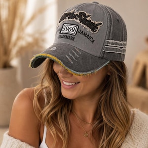 Peut inclure: Une casquette de baseball gris anthracite avec une finition usée et effilochée. L'avant présente un texte brodé "1969 JAMAICA" et "TRADEMARK" avec une écriture cursive. La visière présente un détail effiloché jaune et vert.