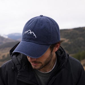 Op de afbeelding: Een marineblauwe baseballpet met een eenvoudig wit geborduurd lijnontwerp van een bergketen op het voorpaneel. De pet heeft een gebogen klep, een knoop aan de bovenkant en gestikte oogjes voor ventilatie, wat zorgt voor een klassieke uitstraling.