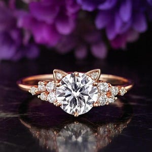 May include: A rose gold ring featuring a large round brilliant-cut centre stone flanked by smaller round accent stones. The setting includes two small triangular points at the top, resembling cat ears, with additional diamonds along the band.