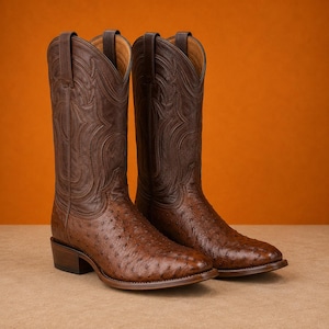 Puede incluir: Un par de botas vaqueras de cuero marrón con un empeine de textura de avestruz y una caña de cuero liso con intrincados patrones de costuras tonales. Las botas tienen una punta clásica, un tacón apilado y tiradores en la parte superior.