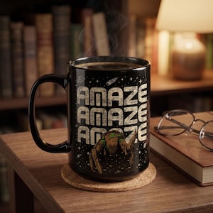 Könnte beinhalten: Ein schwarzer Keramikbecher mit dem wiederholten Text "AMAZE" in einer texturierten, hellen Schriftart. Unter dem Text befindet sich eine Grafik eines braunen, felsigen, mehrbeinigen Wesens vor einem sternenklaren Weltraumhintergrund mit Planeten.