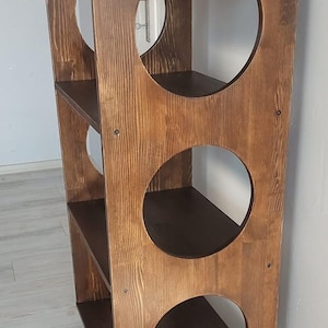 Puede incluir: Una estanter&iacute;a alta de madera de color marr&oacute;n oscuro con tres niveles de estantes horizontales. Los paneles laterales presentan tres grandes aberturas circulares a cada lado y la unidad se apoya sobre cuatro patas de madera c&oacute;nicas.
