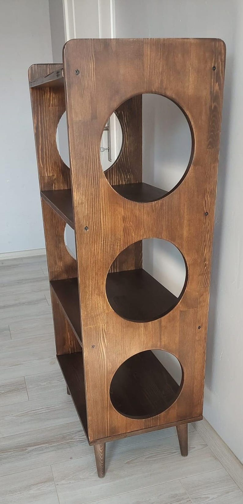 Puede incluir: Una estanter&iacute;a alta de madera de color marr&oacute;n oscuro con tres niveles de estantes horizontales. Los paneles laterales presentan tres grandes aberturas circulares a cada lado y la unidad se apoya sobre cuatro patas de madera c&oacute;nicas.