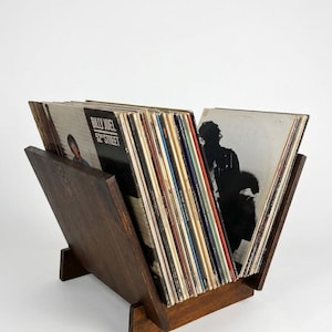 May include: A dark brown wooden V-shaped vinyl record storage rack holding a collection of multiple vinyl records. The front-most record cover features the text Billy Joel 52nd Street, while the other side shows a black and white silhouette.