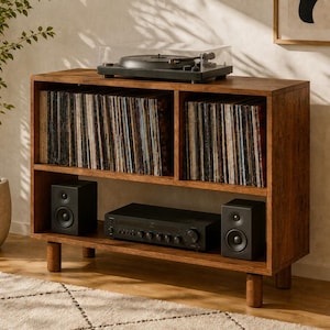 May include: A brown wooden media console with four short legs, featuring two upper cubbies filled with vinyl records and a lower shelf holding a black audio receiver flanked by two black speakers, with a turntable placed on the top surface.