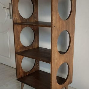 Puede incluir: Una estanter&iacute;a alta de madera de color marr&oacute;n oscuro con tres estantes horizontales y dos paneles laterales verticales. Cada panel lateral tiene tres grandes aberturas circulares y toda la estructura se apoya sobre cuatro patas c&oacute;nicas.