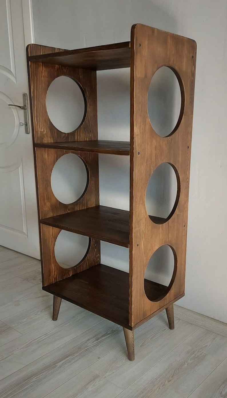 Puede incluir: Una estanter&iacute;a alta de madera de color marr&oacute;n oscuro con tres estantes horizontales y dos paneles laterales verticales. Cada panel lateral tiene tres grandes aberturas circulares y toda la estructura se apoya sobre cuatro patas c&oacute;nicas.