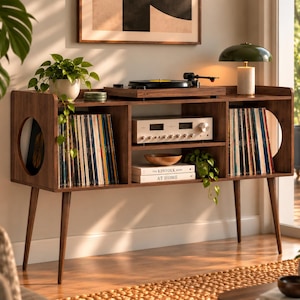 May include: A dark wood media console with four tapered legs, featuring two side compartments with circular cutouts, a central shelf holding a silver audio receiver, and lower shelves containing vinyl records and books titled The Kinfolk At Home.