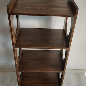 Puede incluir: Una estanter&iacute;a de madera de color marr&oacute;n oscuro con cuatro estantes horizontales y dos paneles laterales verticales. Cada panel lateral presenta dos grandes aberturas circulares. La estructura se apoya sobre cuatro patas cortas y c&oacute;nicas.
