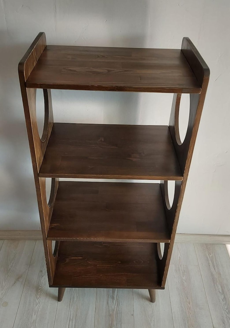 Puede incluir: Una estanter&iacute;a de madera de color marr&oacute;n oscuro con cuatro estantes horizontales y dos paneles laterales verticales. Cada panel lateral presenta dos grandes aberturas circulares. La estructura se apoya sobre cuatro patas cortas y c&oacute;nicas.