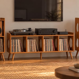 May include: A light brown wooden media console and two matching speaker stands with oval side cutouts. The console holds a black turntable, a black amplifier, and a black streaming device, with vinyl records stored in the lower cubby compartments.