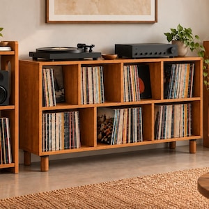 May include: A light brown wooden media console with eight storage cubbies filled with vinyl records, flanked by two matching tall, narrow shelving units. A black turntable and amplifier sit on top of the console, with speakers and plants.