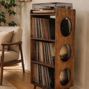 May include: A tall, brown wooden record storage cabinet with three shelves filled with vinyl records. The unit features a top surface holding a black turntable and a side panel with three circular cutouts. It stands on four tapered wooden legs.