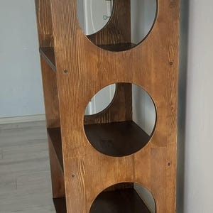 Puede incluir: Una estanter&iacute;a alta de madera marr&oacute;n oscuro que presenta un panel vertical con tres grandes aberturas circulares. La unidad cuenta con cuatro estantes internos y se apoya sobre cuatro patas de madera c&oacute;nicas con veta visible.