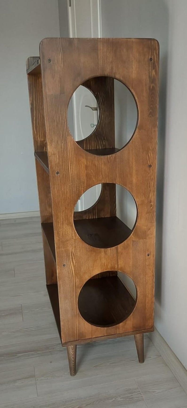 Puede incluir: Una estanter&iacute;a alta de madera marr&oacute;n oscuro que presenta un panel vertical con tres grandes aberturas circulares. La unidad cuenta con cuatro estantes internos y se apoya sobre cuatro patas de madera c&oacute;nicas con veta visible.