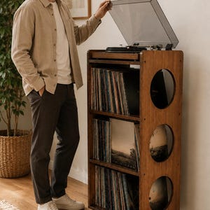 Puede incluir: Un mueble de madera alto y rectangular para guardar discos, con tres estantes abiertos para &aacute;lbumes de vinilo y una superficie superior con un tocadiscos con tapa transparente. El panel lateral tiene tres aberturas circulares.
