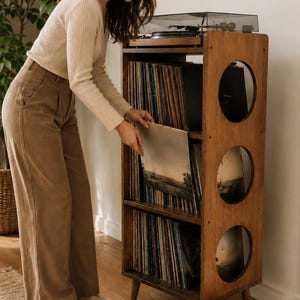 Puede incluir: Un mueble de madera alto y rectangular para guardar discos, con tres aberturas laterales circulares y tres estantes abiertos llenos de vinilos. Un tocadiscos negro se sit&uacute;a sobre la superficie superior del mueble, apoyado en cuatro patas c&oacute;nicas.