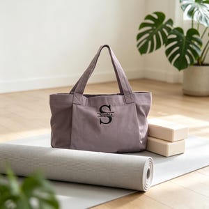 May include: A mauve canvas tote bag featuring a black embroidered monogram with the letter S and the name Sophia centered below it. The bag has two sturdy top handles and a structured rectangular base, positioned near a rolled light gray yoga mat.