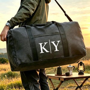 May include: A charcoal gray canvas duffel bag featuring the white letters K and Y separated by a vertical line. The bag includes black nylon webbing handles and an adjustable shoulder strap, designed for carrying personal items during travel.