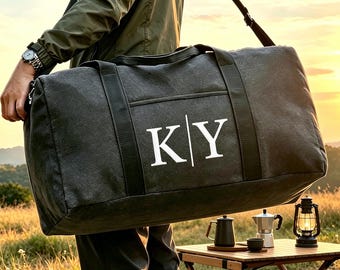 Può includere: Un borsone in tela grigio antracite con le lettere bianche K e Y separate da una linea verticale. La borsa è dotata di manici in nastro di nylon nero e di una tracolla regolabile per il trasporto di oggetti personali.