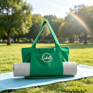 May include: A bright green canvas tote bag featuring two long shoulder straps and a central exterior pocket. The name Isabella is printed in white script inside a white circular outline on the front. A light gray yoga mat is tucked into the pocket.