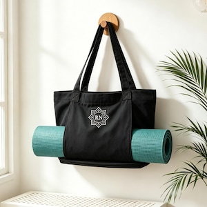 May include: A black canvas tote bag with two long shoulder straps and a teal yoga mat secured horizontally through the front exterior pocket. The bag features a white geometric emblem with the letters RN centered on the front panel.
