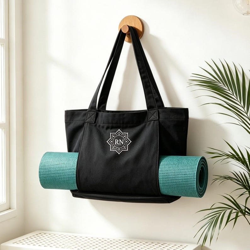 May include: A black canvas tote bag with two long shoulder straps and a teal yoga mat secured horizontally through the front exterior pocket. The bag features a white geometric emblem with the letters RN centered on the front panel.
