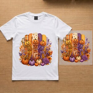 Peut inclure: Un t-shirt blanc avec une impression graphique représentant trois squelettes dansants à tête de citrouille. Les squelettes sont entourés de feuilles d'automne, de fleurs et de citrouilles dans des tons orange, violet et jaune sur un fond rayé.