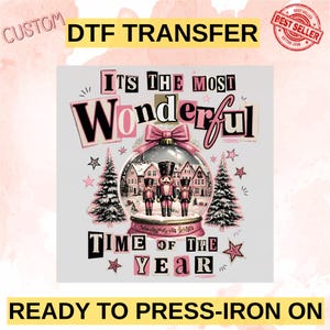 Könnte beinhalten: Ein DTF-Transfer mit einer Schneekugel, in der drei Nussknacker stehen, umgeben von zwei Tannen und Sternen. Der Text lautet ITS THE MOST WONDERFUL TIME OF THE YEAR in schwarzer und rosa Schrift. Das Design enthält eine rosa Schleife oben.