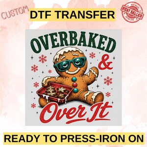May include: A custom DTF transfer featuring a gingerbread person wearing teal sunglasses, holding a red baking tray with star-shaped cookies. The text reads OVERBAKED & OVER IT, surrounded by red snowflakes and stars on a light gray background.