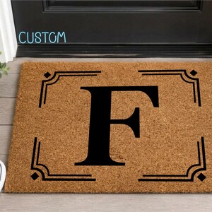 May include: A rectangular tan coir doormat featuring a large black serif letter F in the center, enclosed by a black decorative border with corner accents. The word CUSTOM is printed in light blue text above the doormat.