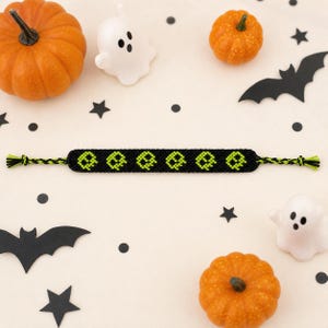 Op de afbeelding: Een zwarte geweven vriendschapsarmband met een herhalend patroon van limoengroene gepixelde spookvormen. De armband heeft gevlochten uiteinden met afwisselend zwarte en limoengroene draden, wat een gedetailleerde geknoopte textuur laat zien.