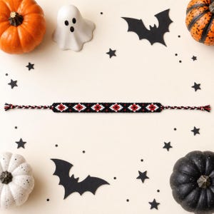 Peut inclure: Un bracelet d'amitié tissé présentant un motif géométrique répétitif en forme de losange avec du fil noir, blanc et rouge. Le bracelet possède des liens tressés aux deux extrémités et est entouré de citrouilles, d'un fantôme et de chauves-souris.