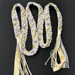 May include: A long braided belt or sash featuring a multi-strand design in pastel shades of lavender, sage green, and cream. The braid is accented with small white faux pearls throughout. The ends feature loose cord tassels and a wide satin ribbon.